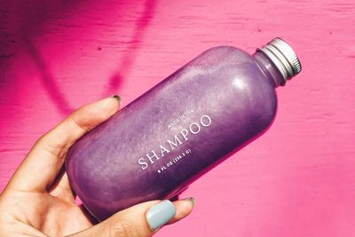 A woman with light blue nails holds a bottle of purple FOB shampoo