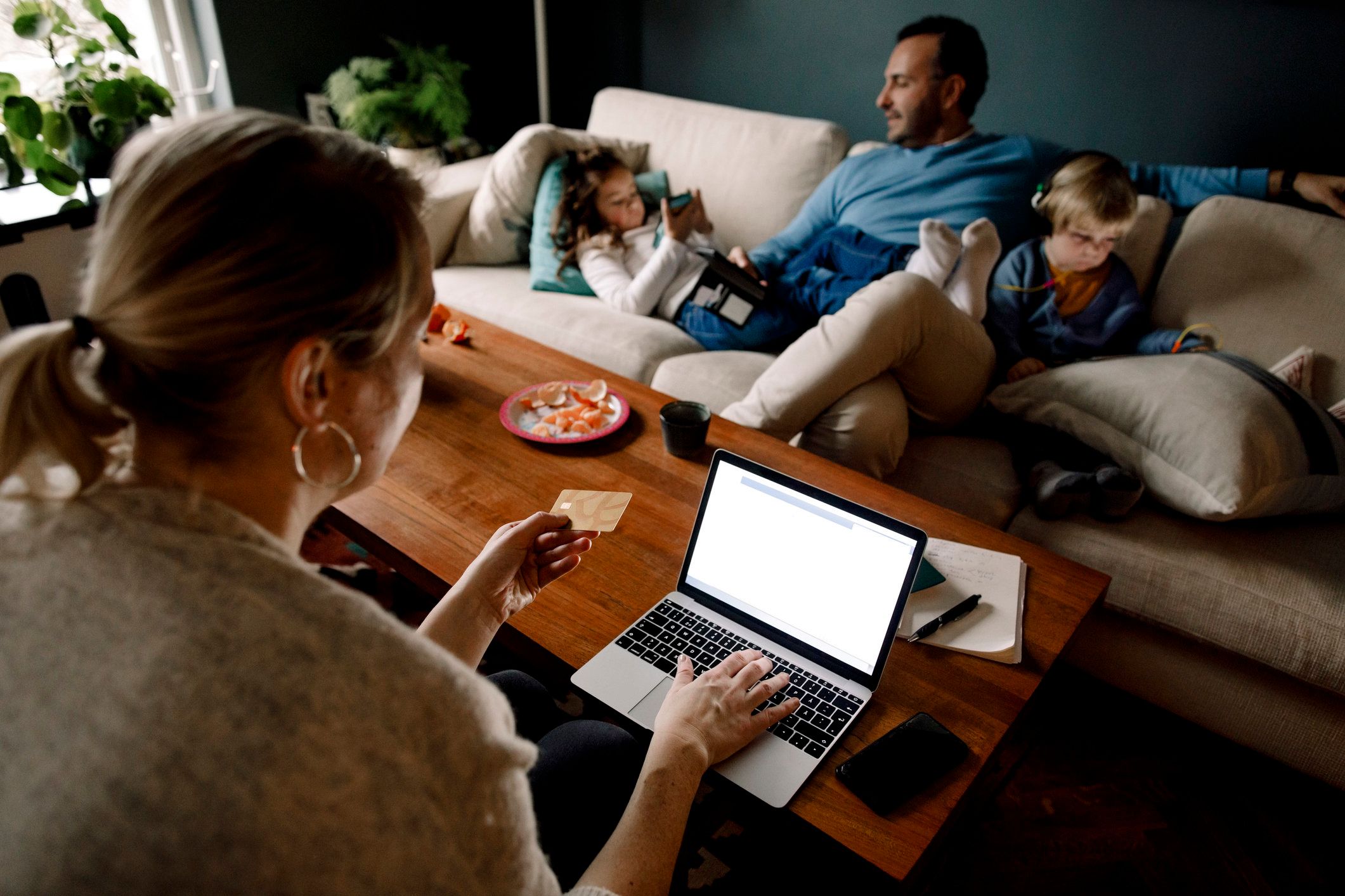 family in a sitting room while mother holds credit card and is on laptop